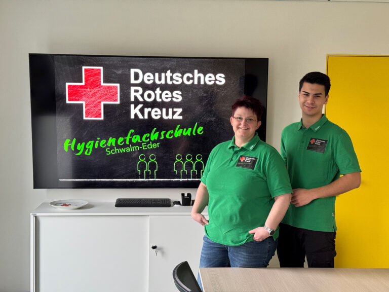 Zwei Dozenten der Hygienefachschule im Schulungsraum vor dem Logo der Hygienefachschule Schwalm-Eder.