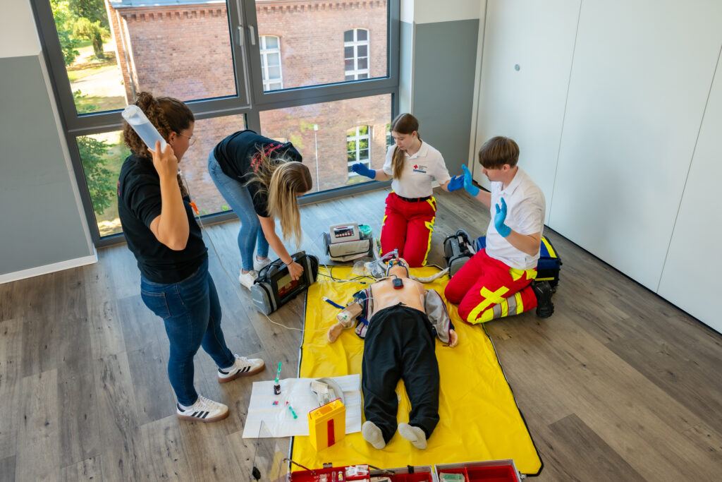 Mehrere Teilnehmer üben gemeinsam die Versorgung einer Reanimationspuppe mit Beatmung, Defibrillator und medizinischer Ausrüstung.