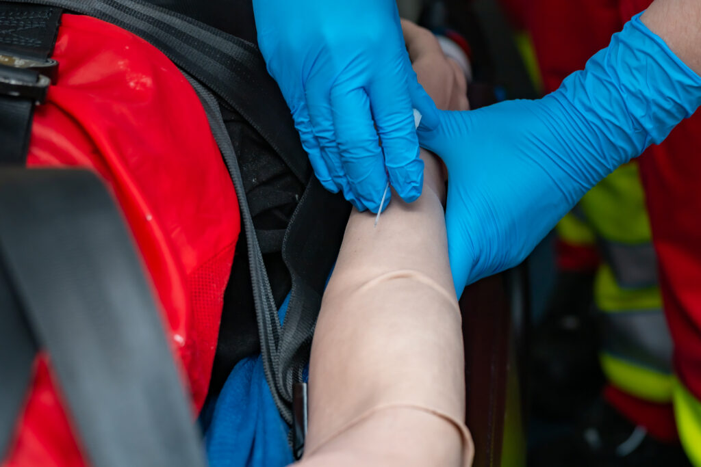 Nahaufnahme einer Venenpunktion an einem Übungsarm mit medizinischer Kanüle und Schutzhandschuhen.