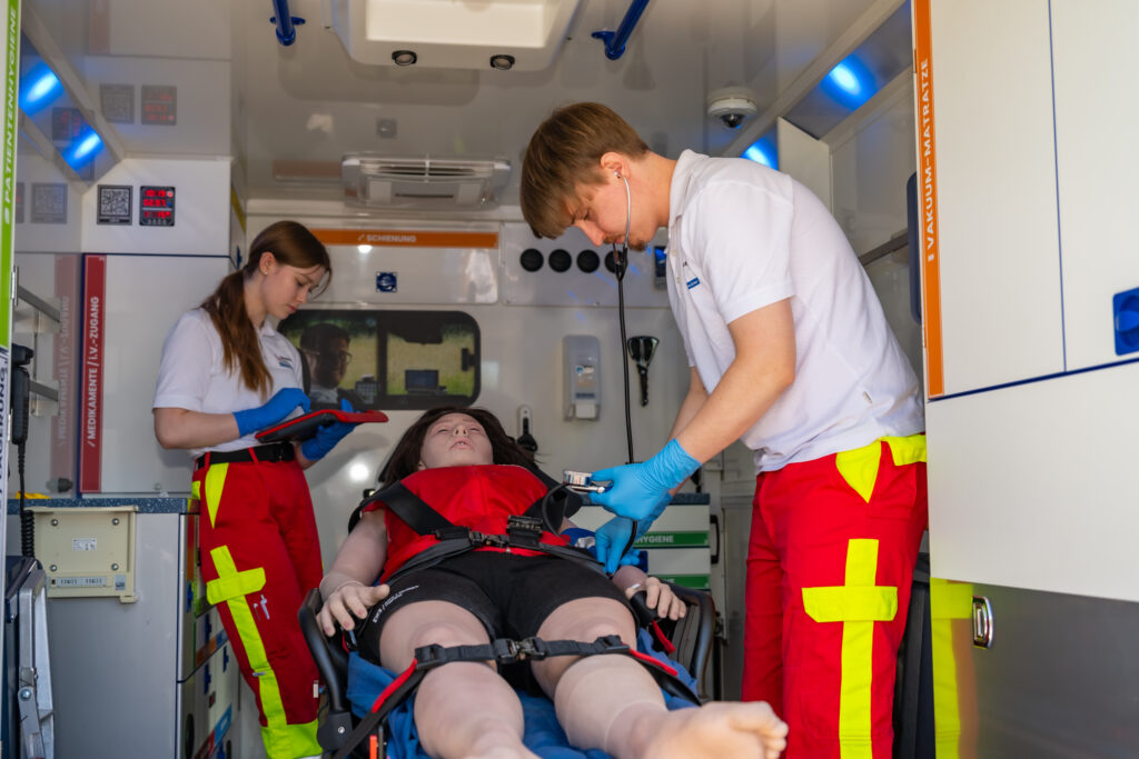 Zwei Teilnehmer untersuchen und dokumentieren den Zustand einer Patientenpuppe auf der Fahrtrage im Rettungswagen.