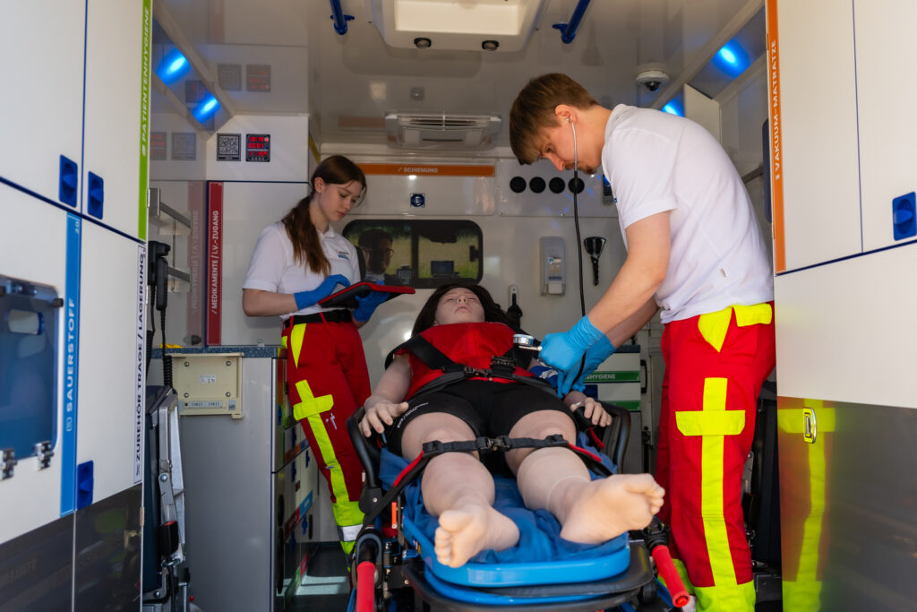 Praktische Rettungsdienstübung im Krankenraum eines Rettungswagens mit Untersuchung und Dokumentation an einer Patientenpuppe.