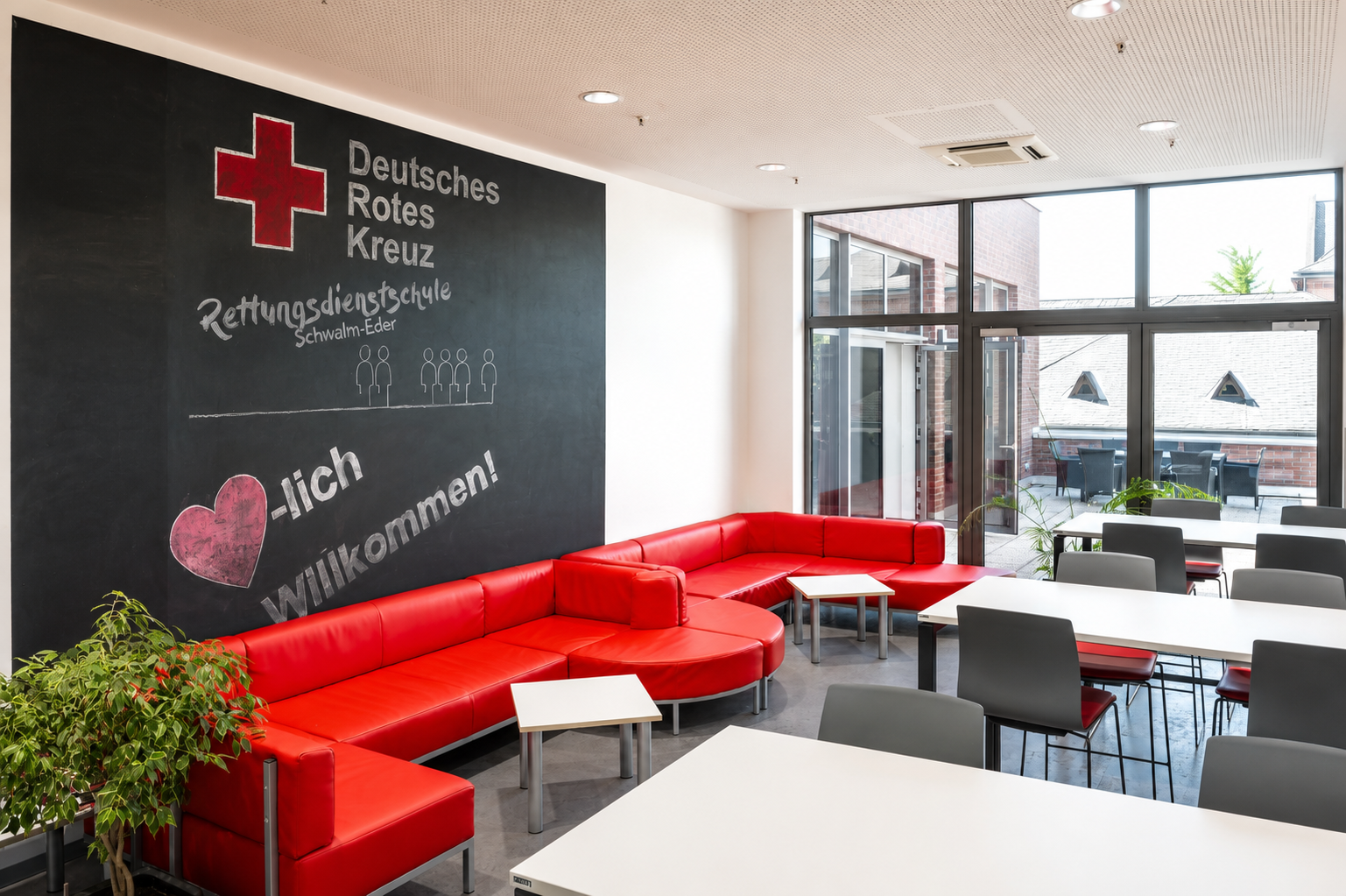 Aufenthaltsraum der Rettungsdienstschule Schwalm-Eder mit roten Sitzmöbeln, Tischen und großer Kreidetafel.